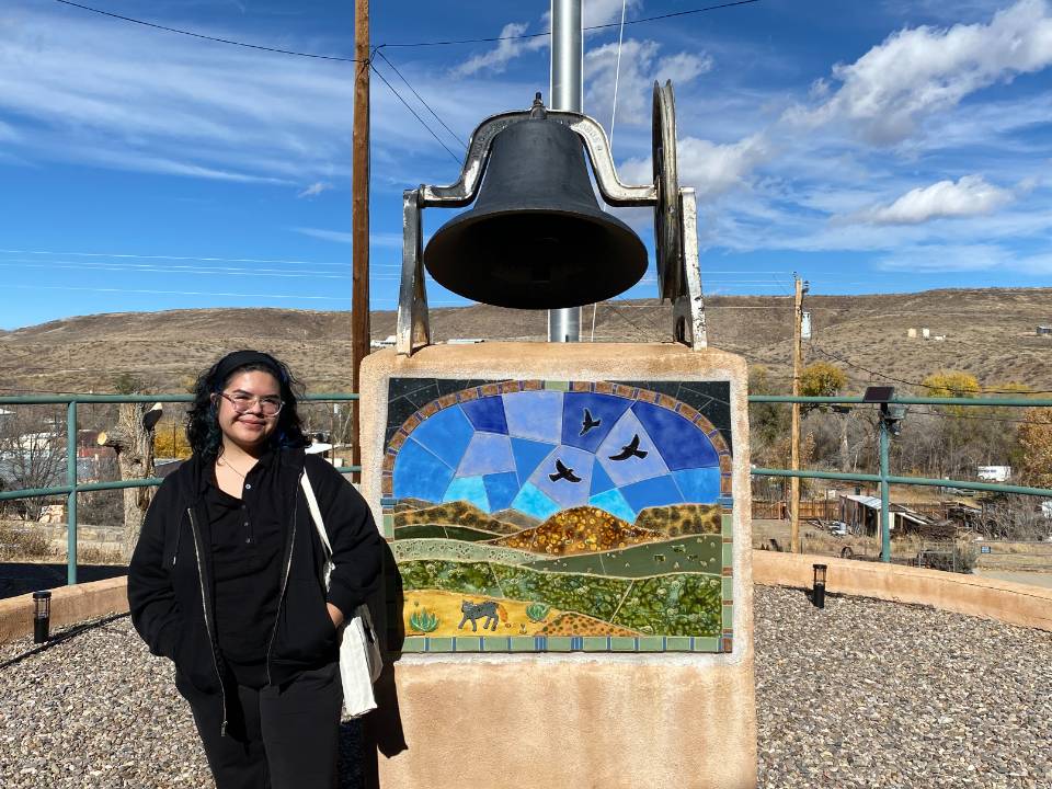 Aspen Coriz-Romero stands in front of a bell with an inlaid image on its base depicting a landscape with hills and birds in the sky at the Hillsboro Community Center and Library in Hillsboro, NM.