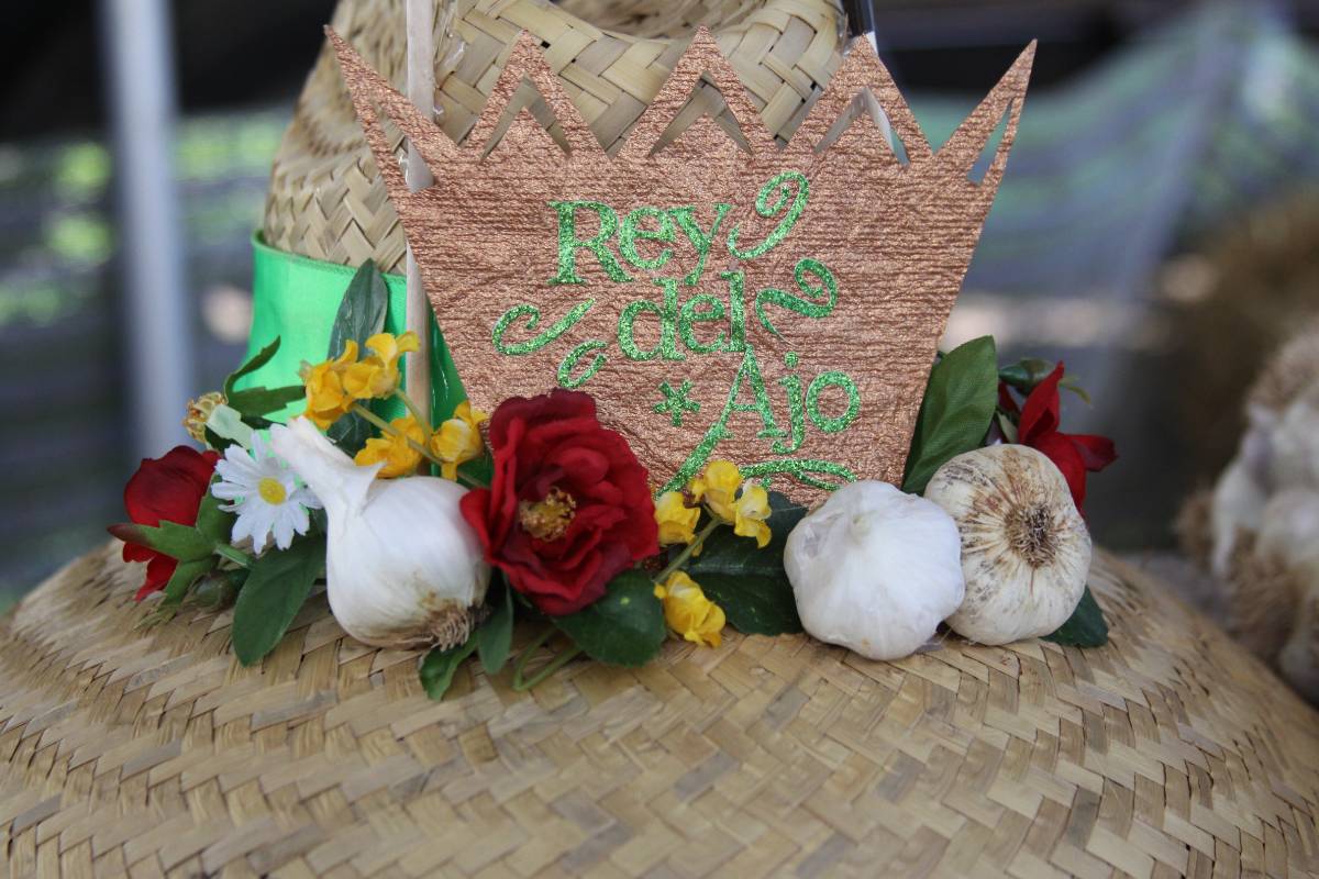 A straw hat with a cut out of a crown, with the words “Rey del Ajo” (King of Garlic) written on it, and a garland of garlic and flowers adorning it. 